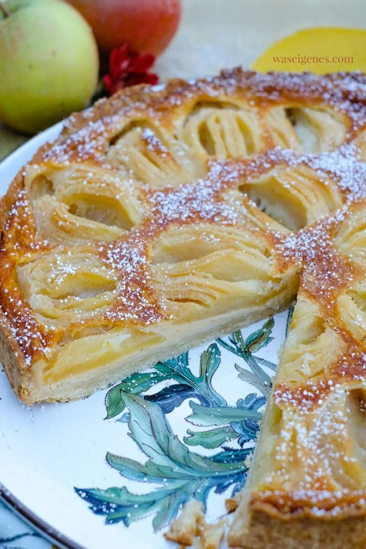 Apfelkuchen mit Sahneguss auf einem Teller, dekoriert mit frischen Äpfeln